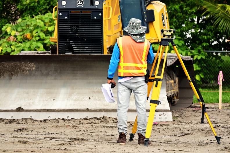 man in yellow and orange jacket and white pants standing beside yellow and black heavy equipment